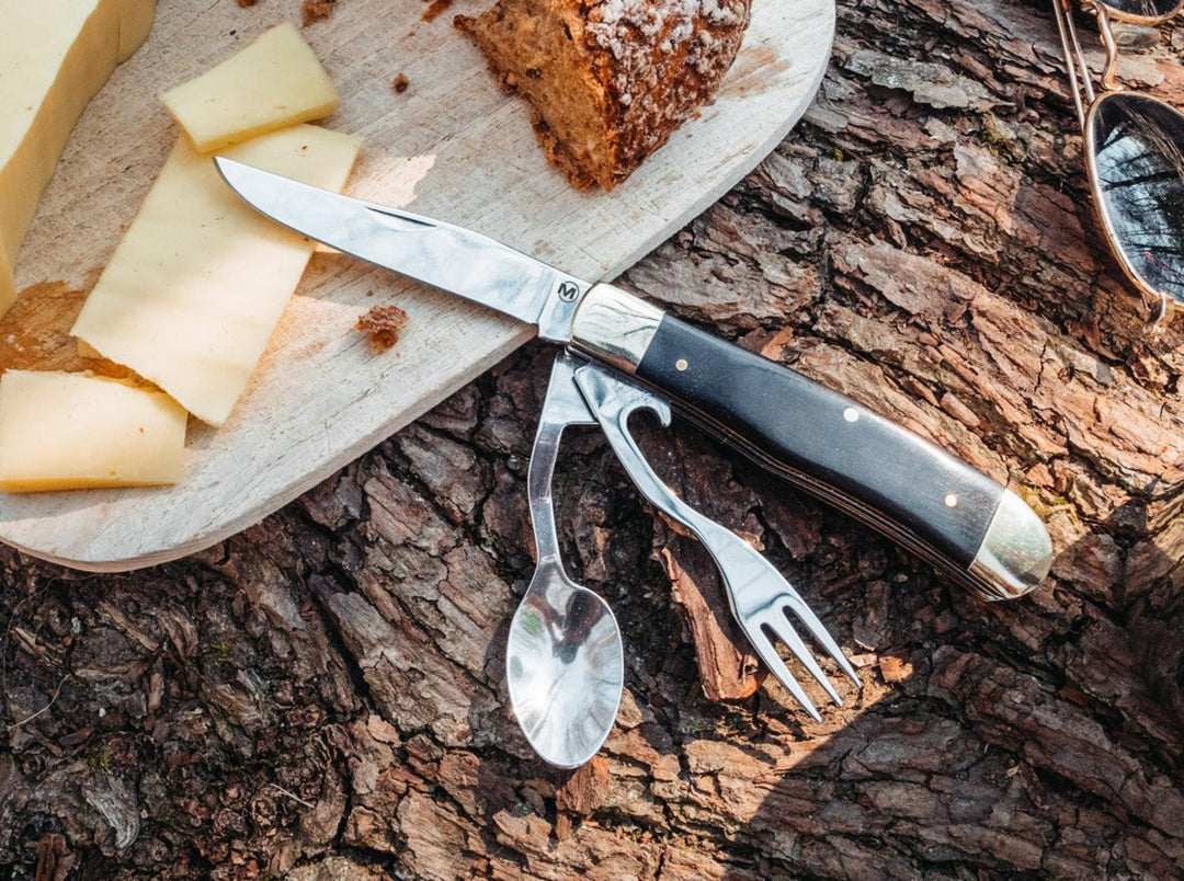 Lifestyle shot of the Böker Magnum Bon Appetite camping cutlery set on tree bark beside cheese and bread on a board