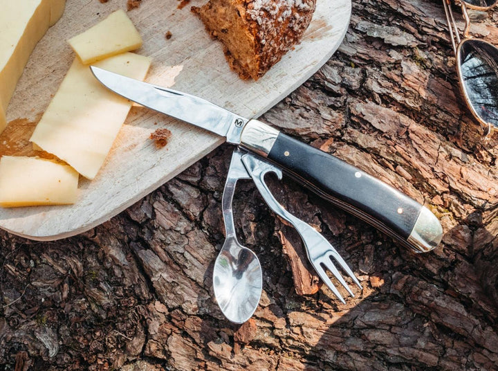 Lifestyle shot of the Böker Magnum Bon Appetite camping cutlery set on tree bark beside cheese and bread on a board