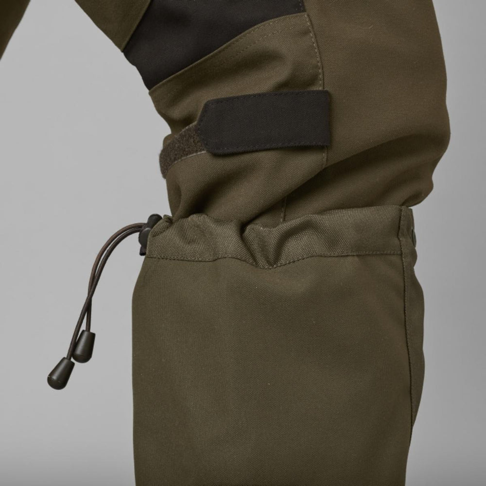 Close-up of the gaiter top fitted over olive trousers on a leg, showing the hook-and-loop tab and elastic drawcord toggle, grey studio background.