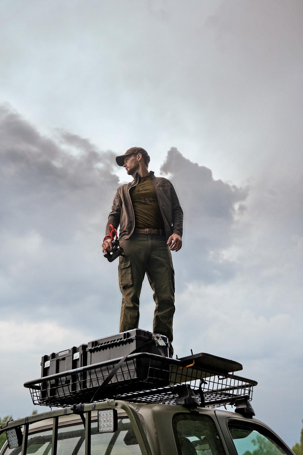 Lifestyle: same stalker photographed from below against a cloudy sky, standing on the pickup roof rack holding binoculars