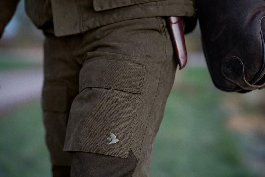 Man walking outdoors wearing the Seeland Helt II trousers, cargo pocket with Seeland duck logo visible, carrying a gun slip