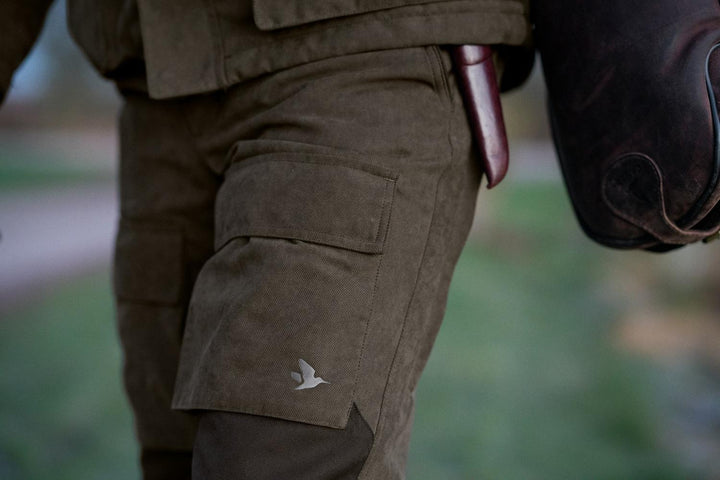 Man walking outdoors wearing the Seeland Helt II trousers, cargo pocket with Seeland duck logo visible, carrying a gun slip