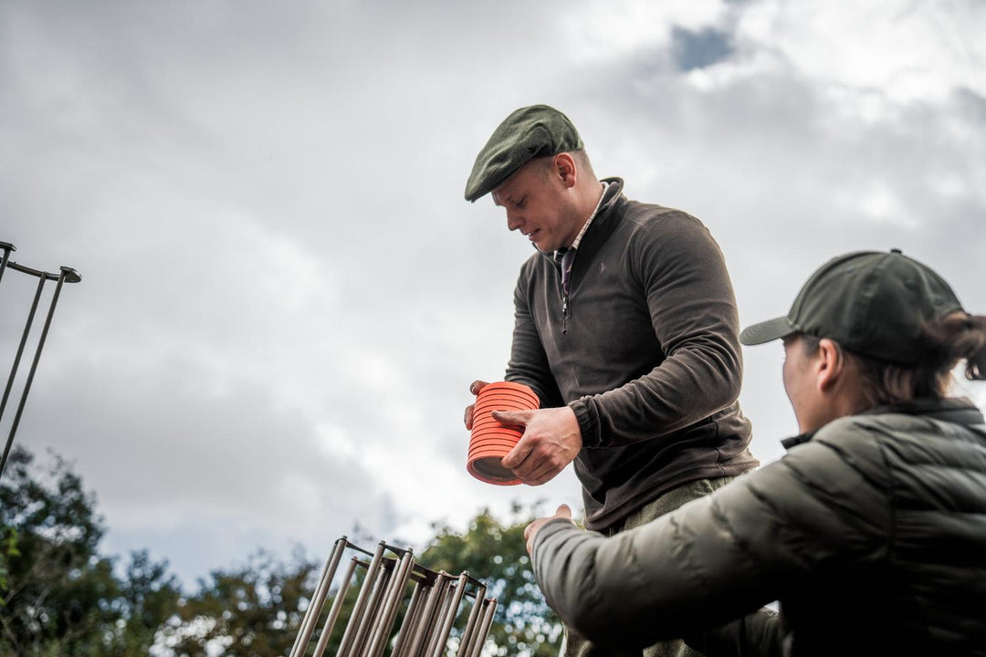 Seeland Benjamin Half Zip Fleece Pine – view 9: lifestyle wide shot of two people at a clay shoot, one wearing the fleece and a flat cap, exchanging orange cartridges
