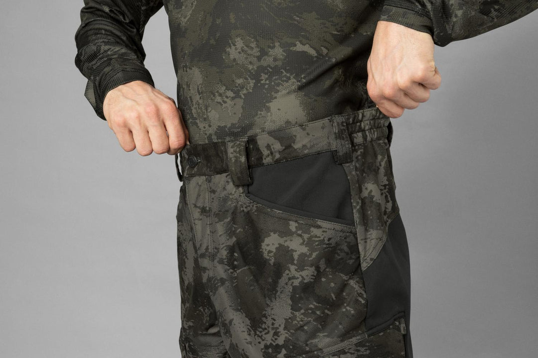 Model detail of the right hip: camo waistband, belt loops and black-trimmed slash pocket, grey studio background