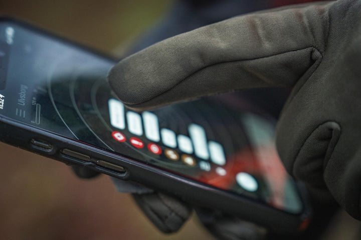 Gloved fingertip tapping a smartphone screen, demonstrating the touchscreen-compatible fingertips on the pine green glove.