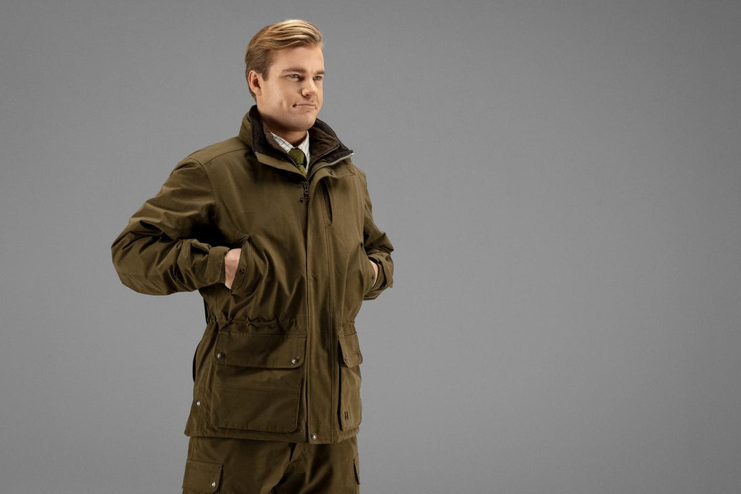 Model wearing the olive Härkila Retrieve jacket over matching trousers, full-length three-quarter view with a hand in the hip pocket