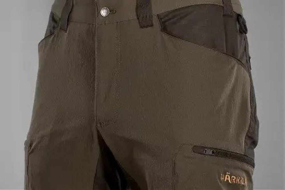 Close-up of the front waistband, button and fly on the olive-green Ragnar trousers, with a zipped thigh pocket and copper Harkila lettering alongside.
