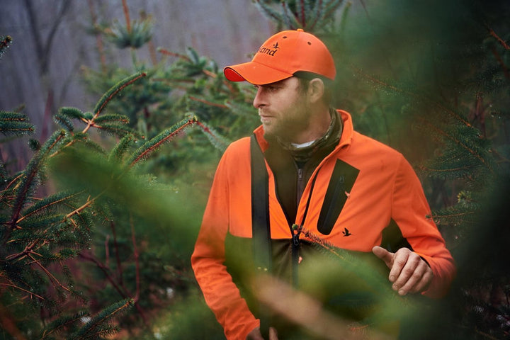 Seeland Force Advanced softshell jacket in blaze orange and dark olive – view 6: lifestyle shot of a hunter wearing the jacket with a matching cap, walking through pine woodland.
