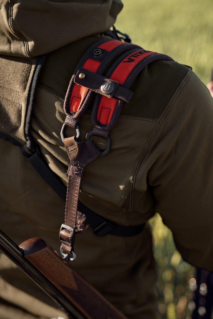 Field shot from behind: a rifle carried on the Härkila Wildboar Pro sling over an olive hooded hunting jacket.