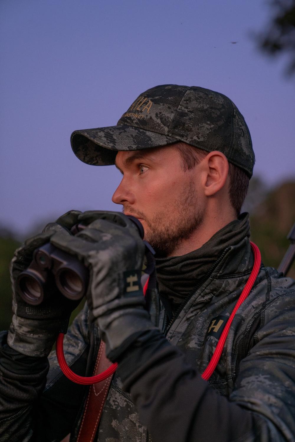 Lifestyle shot at dusk: a hunter in the NOCTYX cap and camo jacket glassing through binoculars under a purple twilight sky.