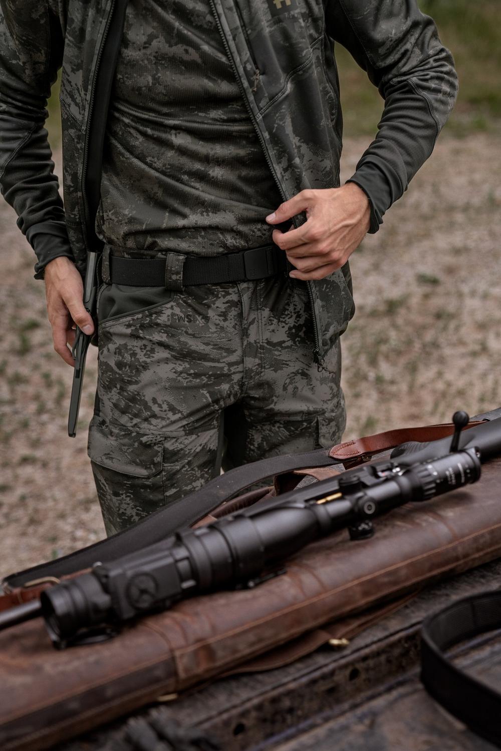 Lifestyle shot: a stalker in camouflage clothing fastening the Harkila Tech belt at the waist, a scoped rifle in a slip lying on the gravel below.