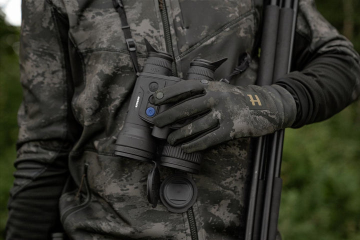 Lifestyle shot of a stalker in NOCTYX camo holding a thermal binocular in gloved hands, rifle slung over the shoulder.