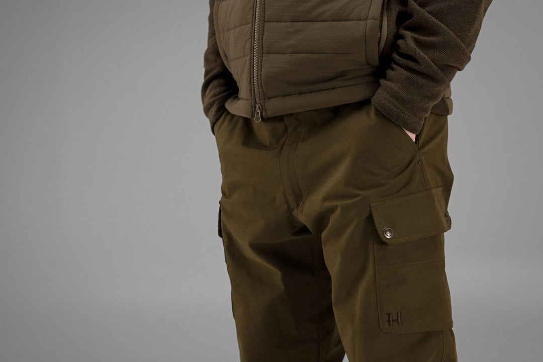 Hip and thigh close-up of the Retrieve trousers worn with a quilted gilet, showing button-flap pocket and Harkila logo