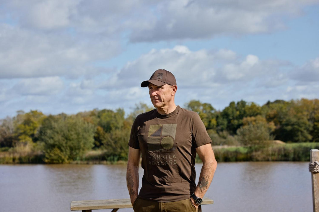 Man wearing the Härkila Anniversary t-shirt and a cap, standing by a lake with trees in the background