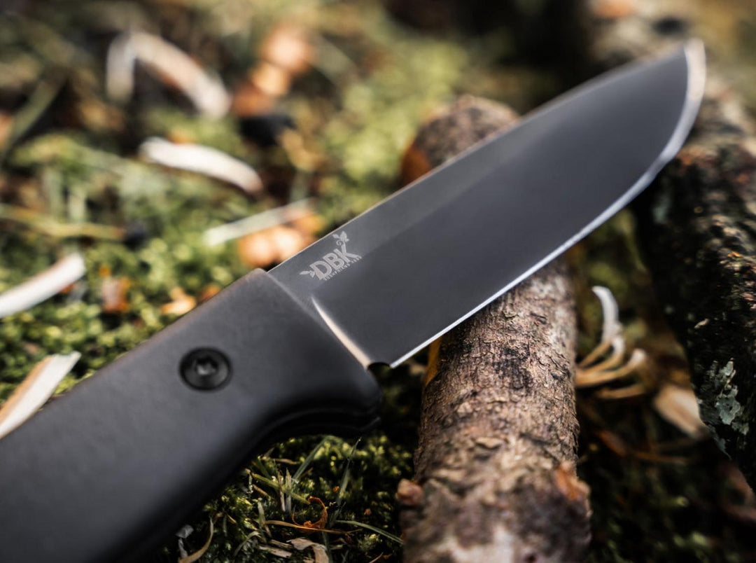 Böker DBK Bushfriend 2000 All-Black blade close-up resting on a log among moss and wood chips, outdoor shot