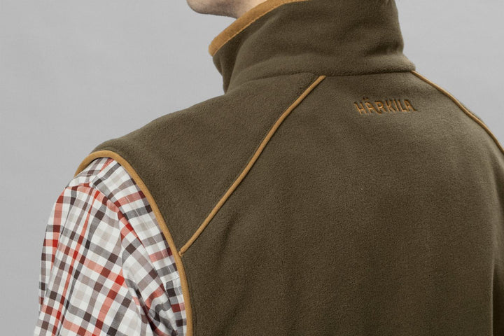 Close-up of the shoulder and collar of the olive Härkila Sandhem 200 waistcoat worn over a check shirt, showing the tan piping and HARKILA embroidery