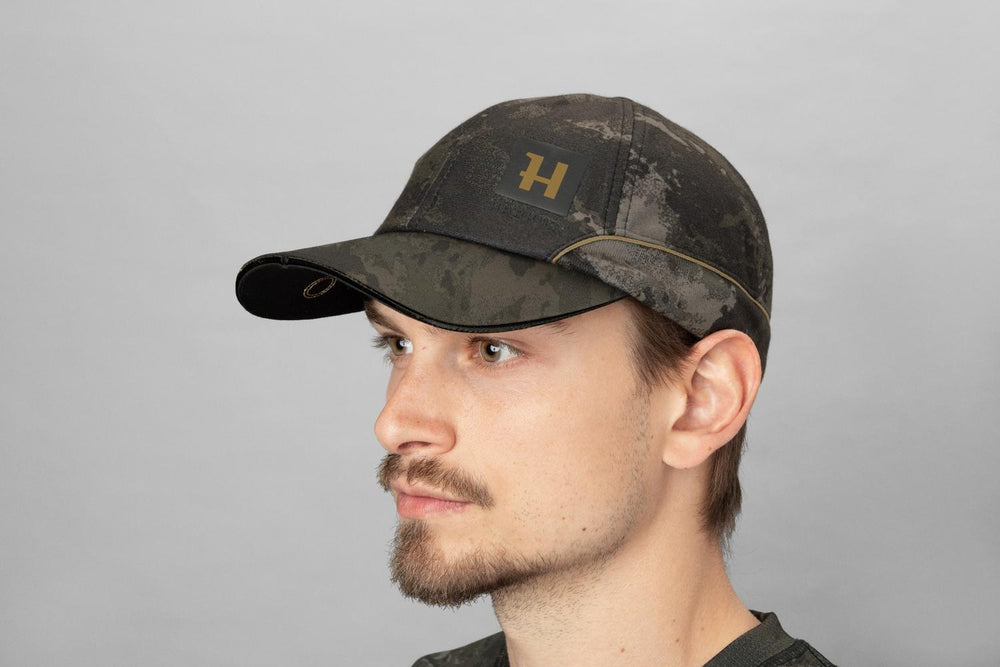 Male model wearing the NOCTYX camo cap, three-quarter front view against a grey studio backdrop.