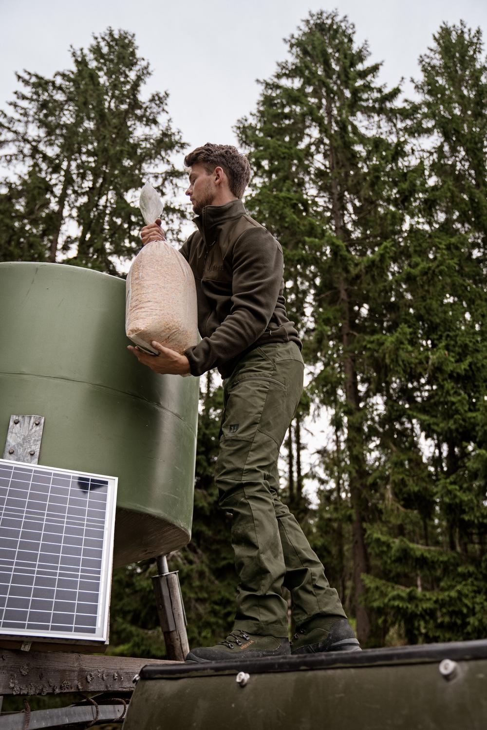 Lifestyle shot: a man in the fleece lifting a sack of feed into a green hopper, conifers and a solar panel behind