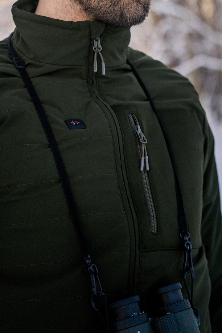 Chest-level shot of a bearded man outdoors in the olive jacket, a binocular strap crossing the zipped chest pocket against a snowy background.