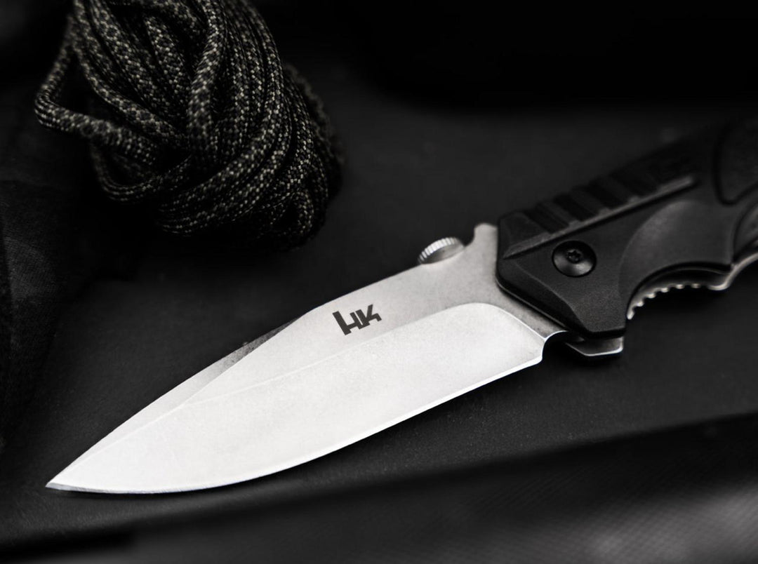 Close-up of the Böker SFP Tactical blade with H&K logo and black handle resting on dark fabric and paracord