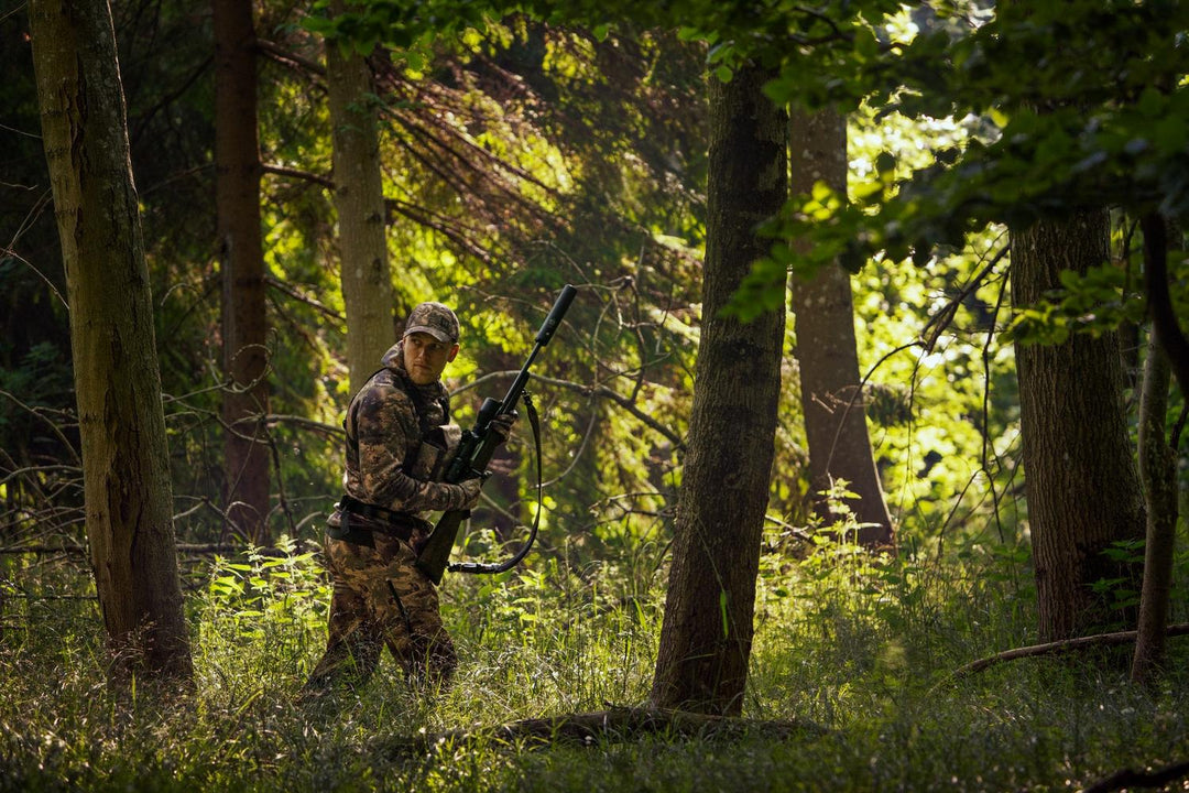 Lifestyle shot: a stalker in Härkila AXIS MSP Forest camo carrying a rifle through sunlit summer woodland