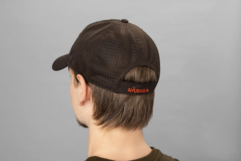 Model wearing the Härkila Impact cap, rear view showing the back panel and the orange HÄRKILA logo across the closure strap.