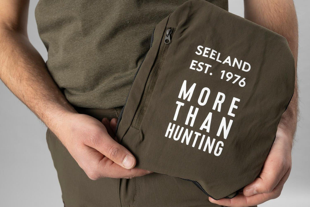 Close crop of hands folding the zip-off leg sections into the olive Seeland 'Est. 1976 More Than Hunting' stuff pouch