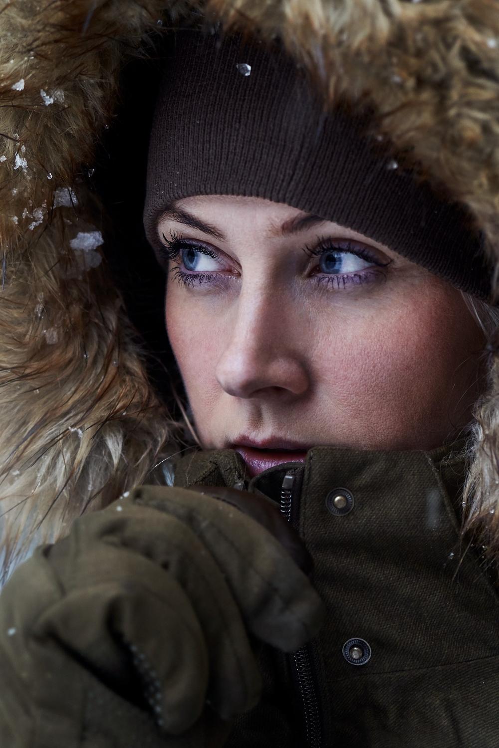 Close-up portrait of a woman in a brown beanie and the fur-trimmed olive hood, gloved hand raised, looking to the side