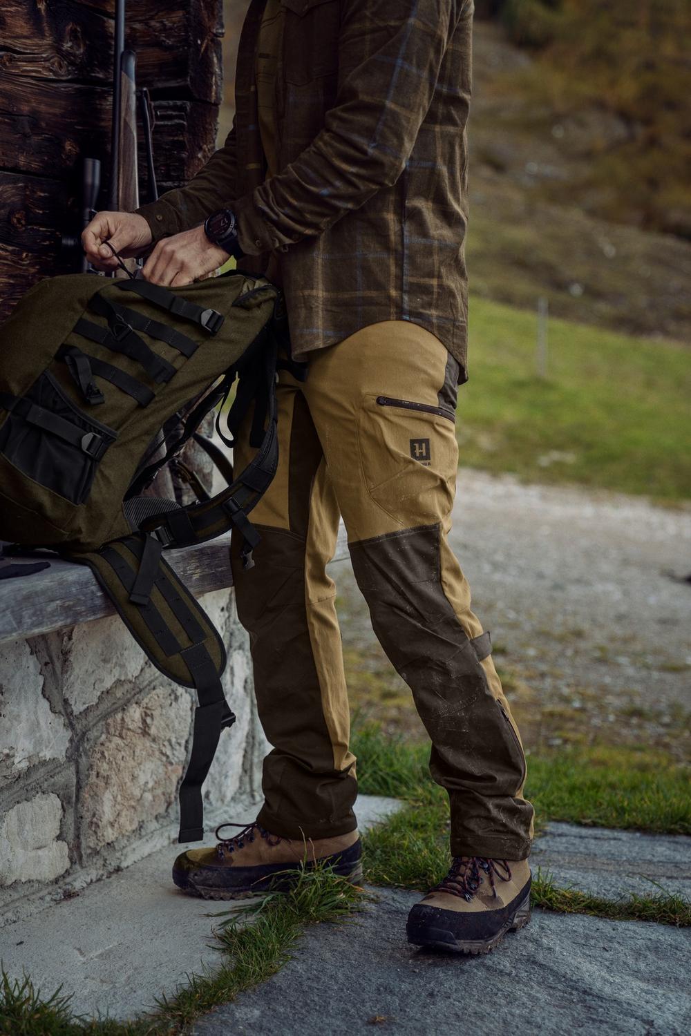 Lifestyle shot: a stalker in Härkila Ragnar trousers lifts a rucksack from a stone wall on a hill track, shown from the chest down