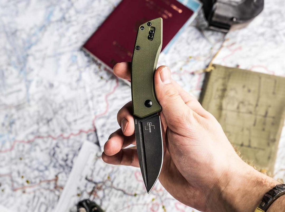 Hand holding the olive green Böker Plus Worldwide 2.0 folding knife open over a map and passport, lifestyle shot