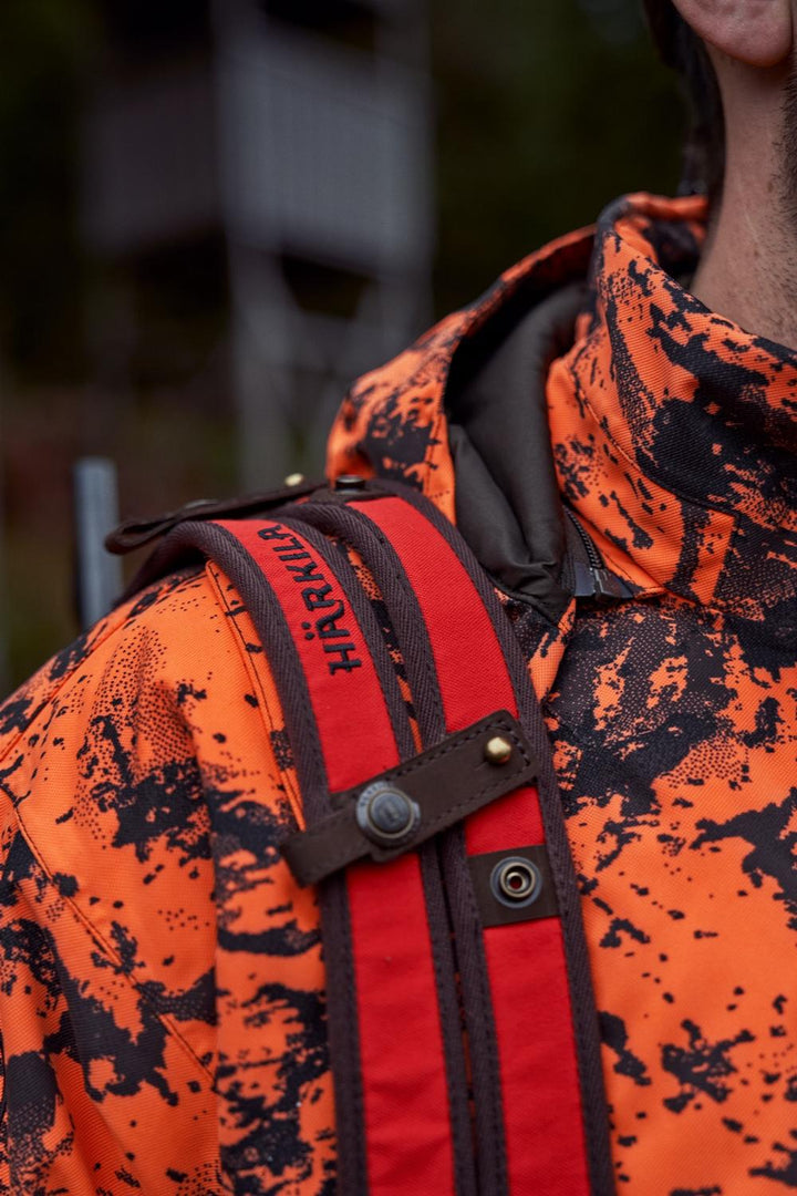 Close-up of the Härkila Wildboar Pro sling on the chest, showing the orange webbing, brown trim, Härkila lettering and press-stud adjusters.