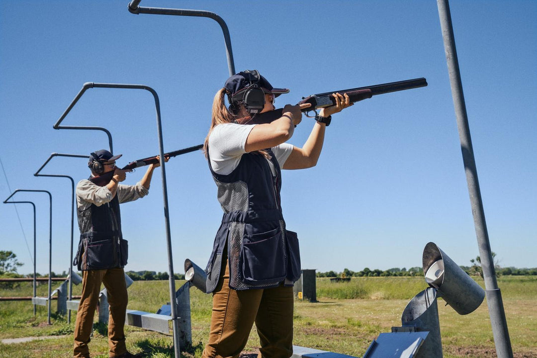 Lifestyle shot: a woman in the Seeland Skeet II Lady waistcoat shooting an over-and-under shotgun on a skeet layout, a second shooter behind her