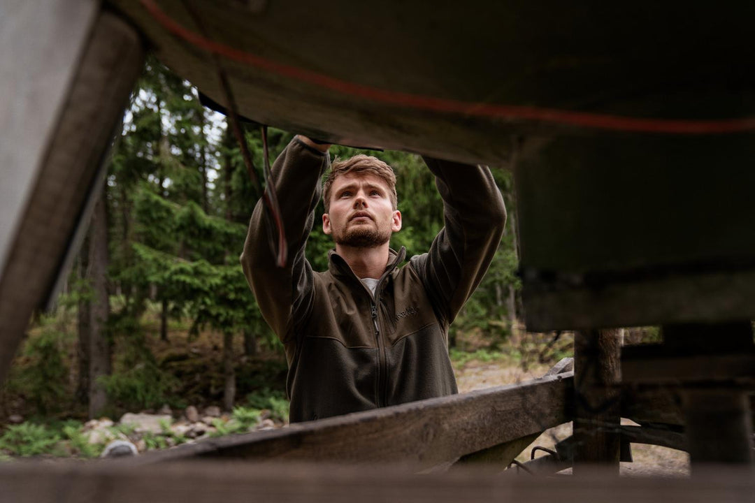Lifestyle shot: a man in the olive fleece reaching up under a raised feeder hopper in woodland