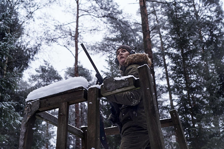 Lifestyle: woman in the fur-hooded jacket lifting a rifle onto the rail of a snow-covered high seat