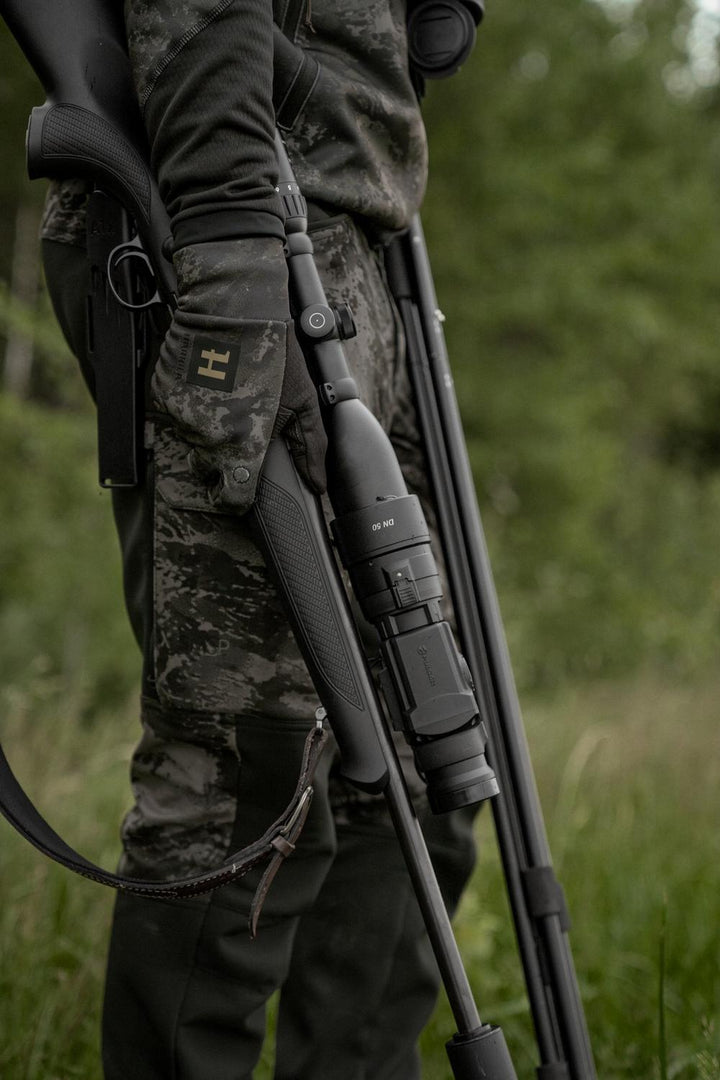 Field shot: stalker wearing the NOCTYX trousers, carrying a scoped rifle and shooting sticks against summer greenery