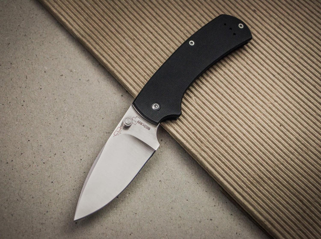 Böker Plus XS Drop folding knife with black handle and satin blade open on a grey and wood surface, lifestyle shot