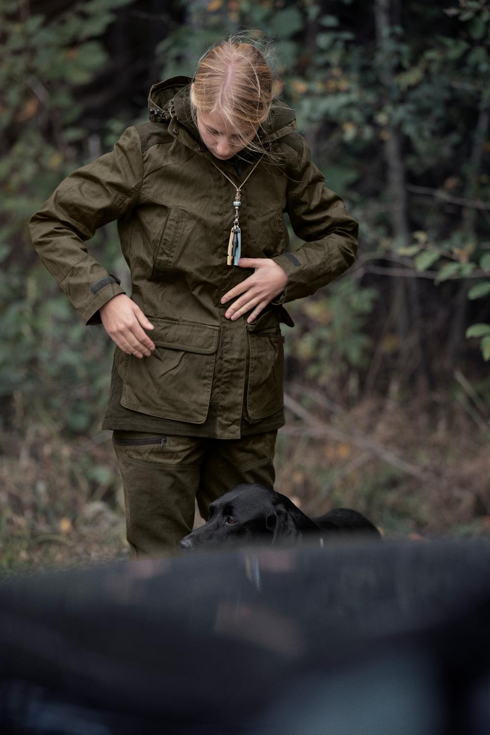 Lifestyle shot: a woman in the olive Seeland Key-Point jacket and trousers standing in scrub, adjusting the jacket, viewed head to knee.