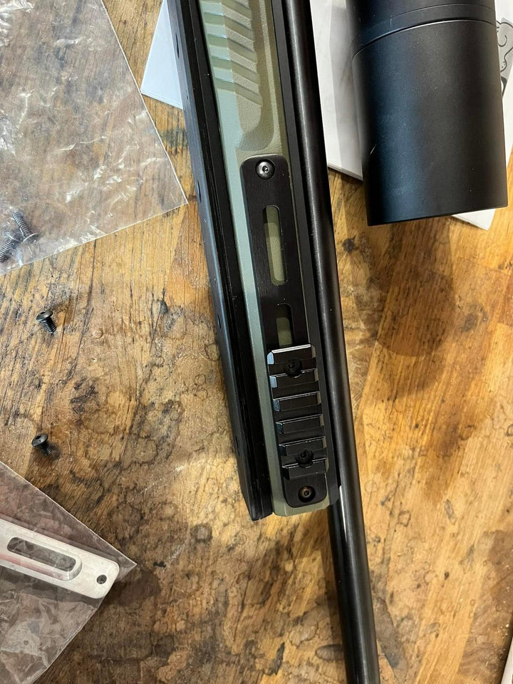 Nieload rail fitted to a bolt-action rifle forend, the rifle stood on a wooden workbench with a sound moderator and tools behind