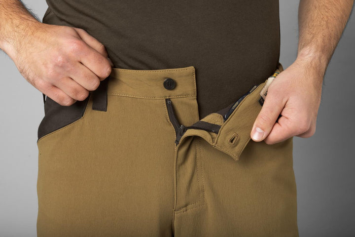 Close-up of a hand opening the button-and-zip fly on the Härkila Ragnar trousers, khaki front with dark brown hip panel