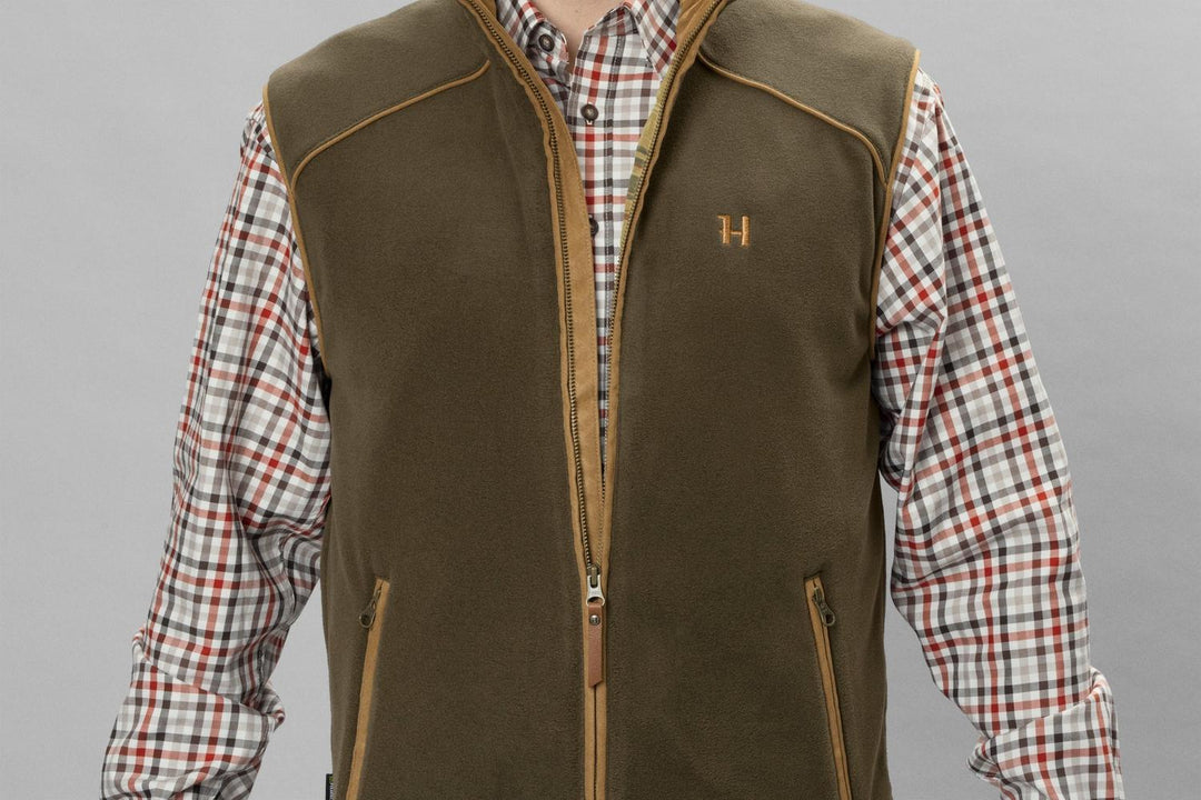 Close-up of the olive Härkila Sandhem 200 waistcoat worn open over a check shirt, showing the zipped hand pocket and tan-piped armhole