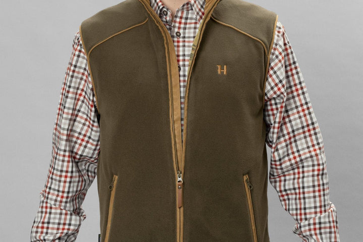 Close-up of the olive Härkila Sandhem 200 waistcoat worn open over a check shirt, showing the zipped hand pocket and tan-piped armhole