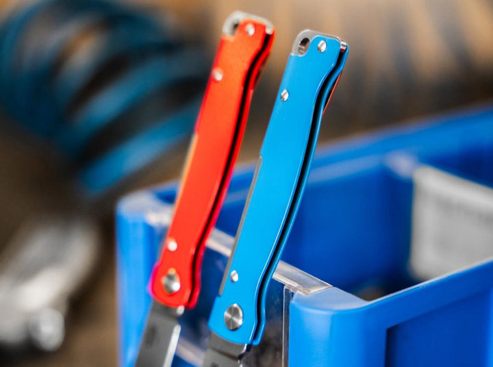 Red and blue Böker Plus Atlas knives standing upright in a blue parts bin on a workbench, lifestyle shot