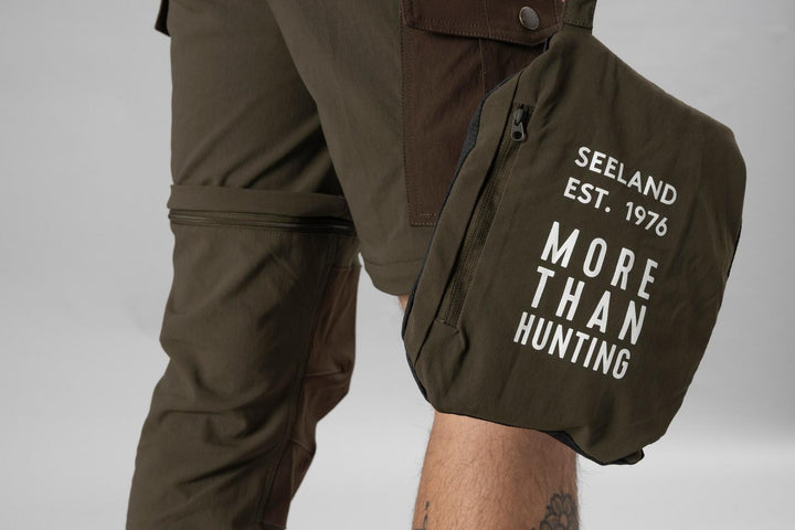 Model standing in the Seeland Birch trousers as shorts, holding the removed lower legs stowed in the Seeland 'More Than Hunting' pouch