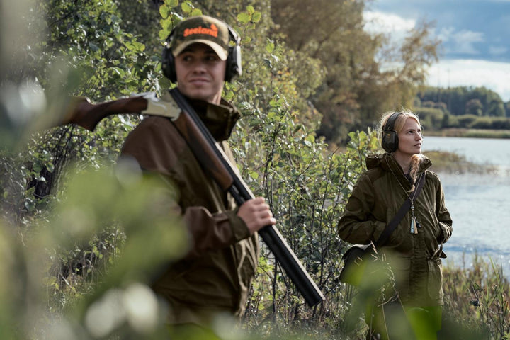 Lifestyle shot by a lake: a shooter in a cap carrying a broken shotgun over the shoulder while a second person in olive stands in the bushes behind.
