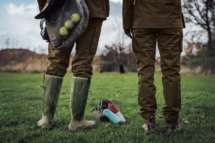 Lifestyle: two people in Harkila Retrieve trousers and wellingtons on a field, dummy bag and training dummies at their feet