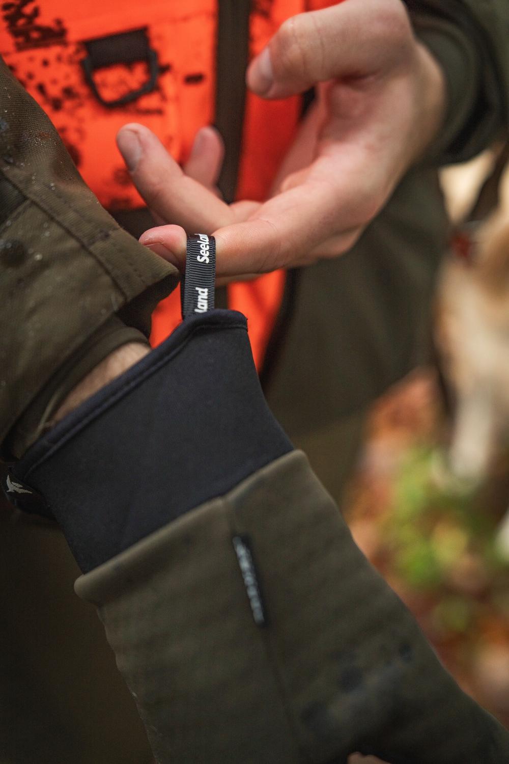 Hunter easing the Seeland glove on over the black neoprene cuff, bare hand and blaze orange vest behind, woodland out of focus.
