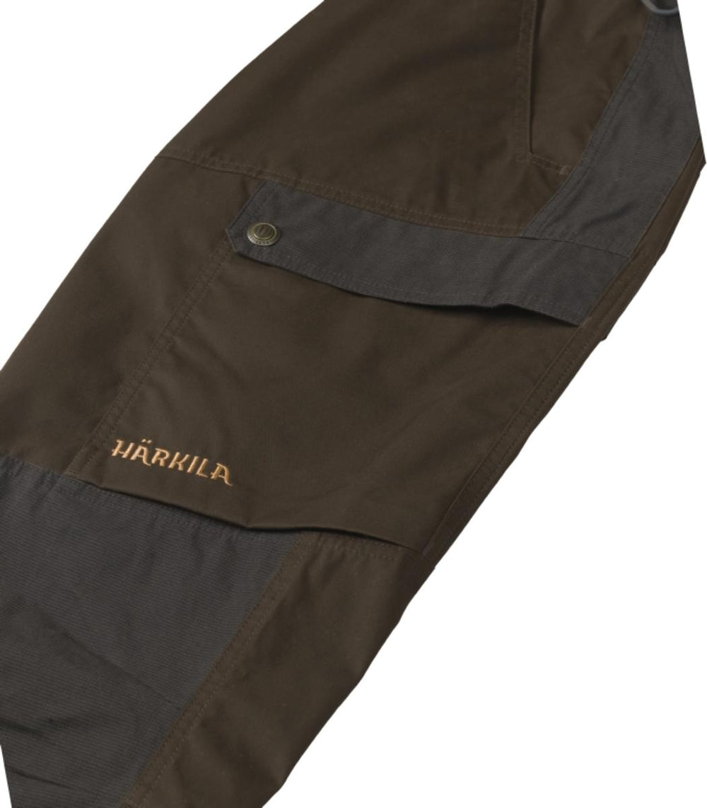 Close-up of the Harkila Asmund trousers thigh cargo pocket: button-down flap over olive-brown fabric with the orange embroidered HARKILA logo and a charcoal reinforcement panel below.