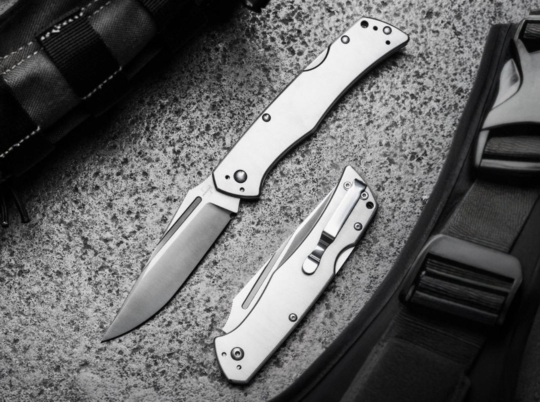 Böker Plus MEK folding knife open beside a closed second knife on a textured stone surface and bag strap