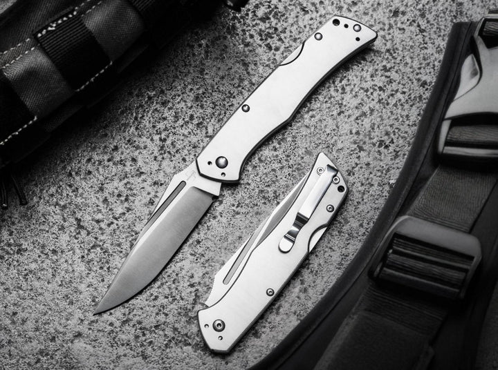 Böker Plus MEK folding knife open beside a closed second knife on a textured stone surface and bag strap