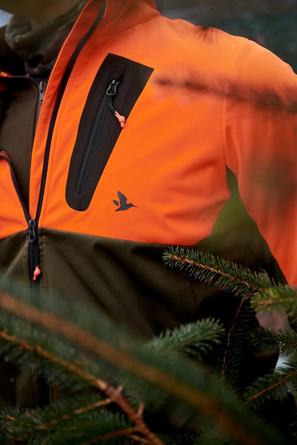 Seeland Force Advanced softshell jacket in blaze orange and dark olive – view 7: close-up of the chest zip pocket and shoulder seam, shot outdoors against pine branches.